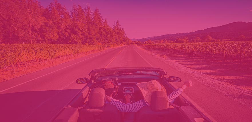 Couple in convertible car driving through vineyards with forests and mountains in the distance.