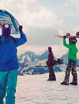 Three snowboarders holding their boards look out at snow covered mountain views.