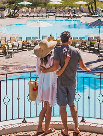 Couple at hotel resort spa, looking out over balcony at two pools and the ocean.